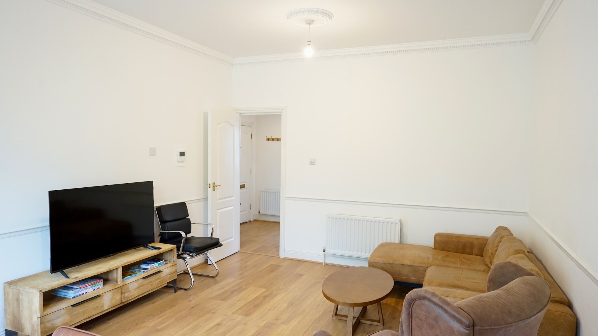 Un salon avec un téléviseur à écran plat posé sur un meuble en bois, un fauteuil de bureau noir, un canapé marron, une petite table ronde et un radiateur.