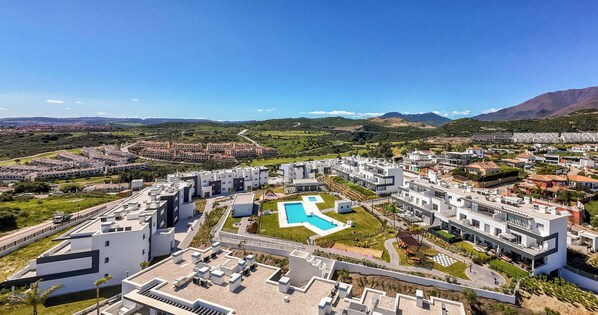 A modern residential complex with a central swimming pool, surrounded by greenery and mountains in the background. - Valle Romano | Pool - terrace - gym (Estepona)