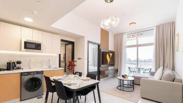 Una cocina moderna con microondas, cafetera y lavadora; una zona de comedor con una mesa preparada para dos; una sala de estar con sofá y vistas a la ciudad.