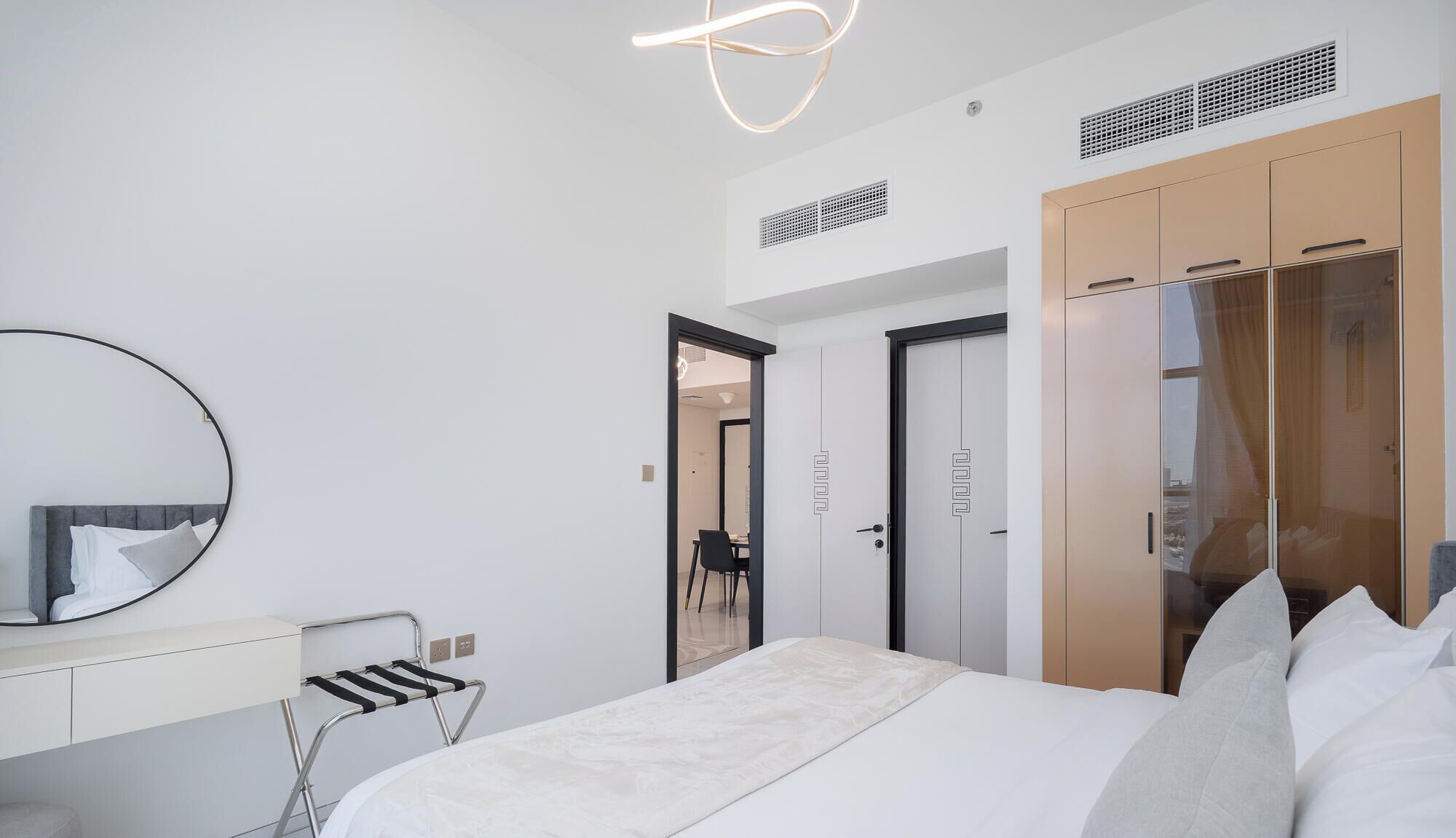 Une chambre d’hôtel moderne avec un lit, un miroir rond, une armoire et une salle de bain visible à travers une porte ouverte.