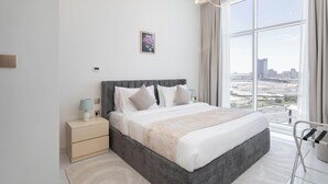 A modern bedroom with a large bed, a nightstand, a bedside table, a wall-mounted light fixture, and a city view through large windows. - Vogue | Skyz by Danube near Dubai Miracle Garden (Dubai)