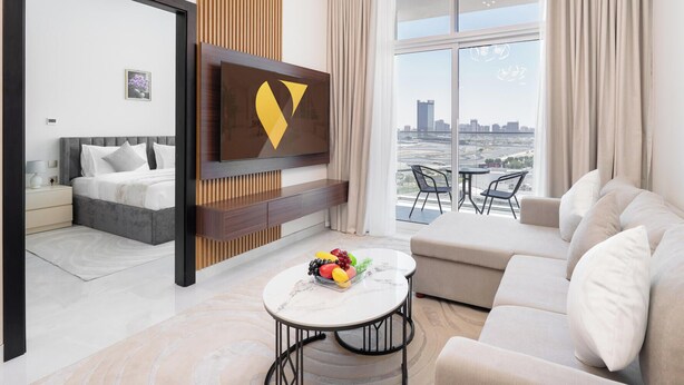 A modern hotel room with a bed, a sofa, a dining table with fruit, a TV, and a city view. - Vogue | Skyz by Danube near Dubai Miracle Garden (Dubai)