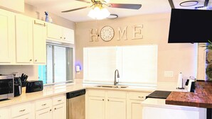 A kitchen with white cabinets, a ceiling fan, a 'HOME' sign, a microwave, and a trash can.