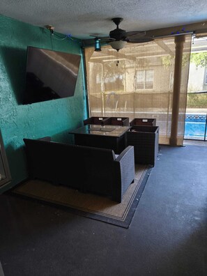 Una habitación con ventilador de techo, televisor instalado en la pared y un amplio ventanal con vista a una piscina.