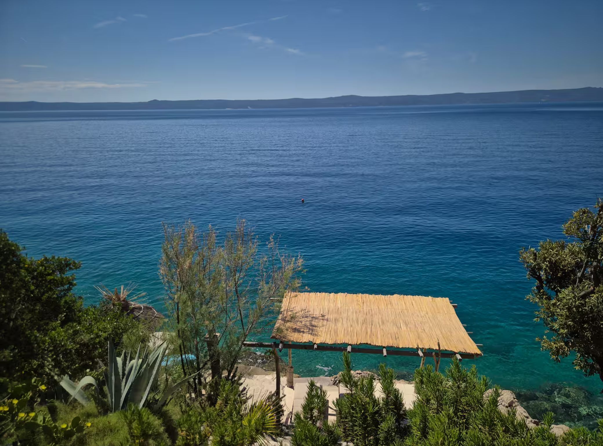 A beach view with clear blue water, a thatched-roof structure, and surrounding greenery.
