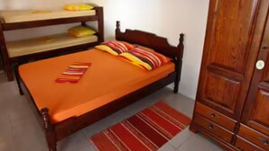 Uma cama de solteiro com estrutura de madeira, um travesseiro listrado em vermelho e amarelo, e um tapete listrado em vermelho e branco no chão.