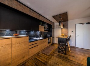 A modern kitchen with wooden cabinets, a black countertop, and a dining area with a chair. - Deluxe Apartment Bono (Sinj)