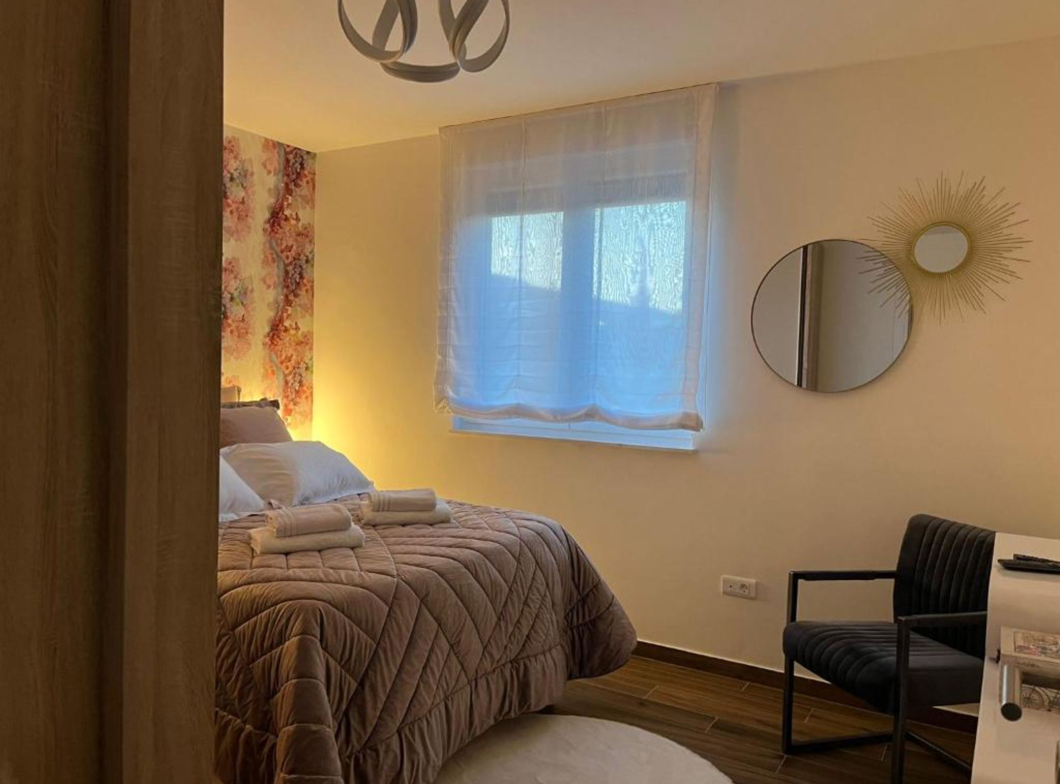 Un dormitorio con una cama, una silla, un ventanal con cortinas, un espejo montado en la pared y un tapiz floral.