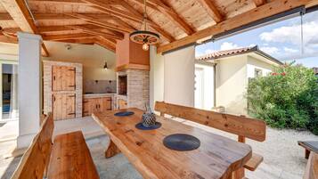 Un espace de repas extérieur en bois, avec des bancs et une table, une cheminée en briques et une vue sur une maison et la verdure.