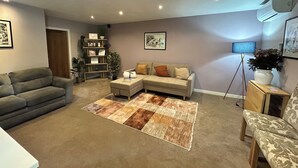 A living room with a sofa, a coffee table, a floor lamp, a bookshelf, and a potted plant. - The Lodge (Brighouse)
