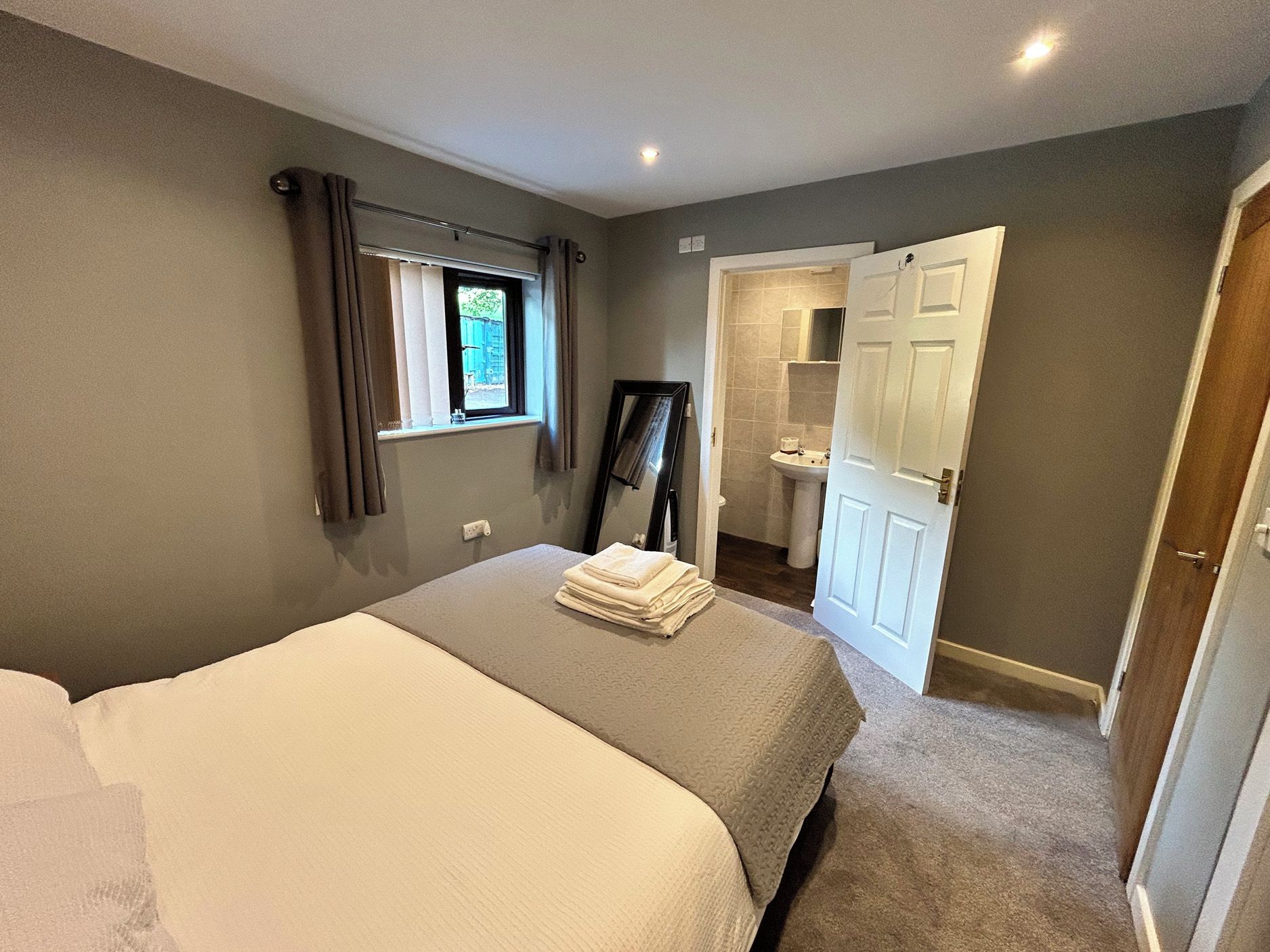 A bedroom with a bed, a window with curtains, a mirror, and a bathroom visible through an open door.