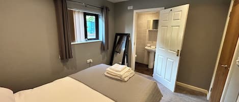 A bedroom with a bed, a window with curtains, a mirror, and a bathroom visible through an open door.