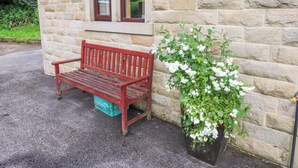 A stone building with a red bench and a planter with white flowers. - The Lodge (Brighouse)