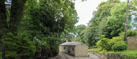 A small stone building with a sloped roof, surrounded by dense greenery and a paved path leading to it.