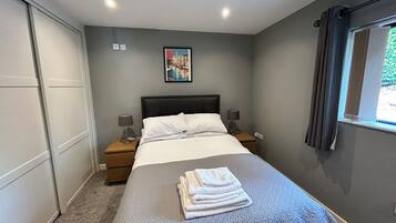 Ein ordentlich bezogenes Bett mit einem gefalteten Handtuch, ein Nachttisch mit einer Lampe und ein gerahmtes Bild an der Wand.