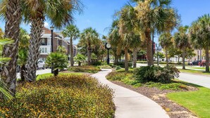 A paved pathway flanked by palm trees and flowering plants, leading to a building with a brick facade and a clock. - Riviera Suites | Near Beach & Boardwalk (Myrtle Beach)
