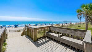 Eine Holzterrasse führt zu einem Strand mit einer Bank, einer Palme und Sonnenschirmen im Hintergrund.