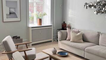 En stue med sofa, to lænestole, sofabord, radiator, et vindue med udsigt og en vægmonteret lampe.