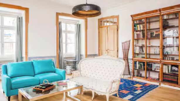 Ein Wohnzimmer mit einem türkisfarbenen Sofa, einem weißen Sessel, einem Holztisch und einem großen Bücherregal.