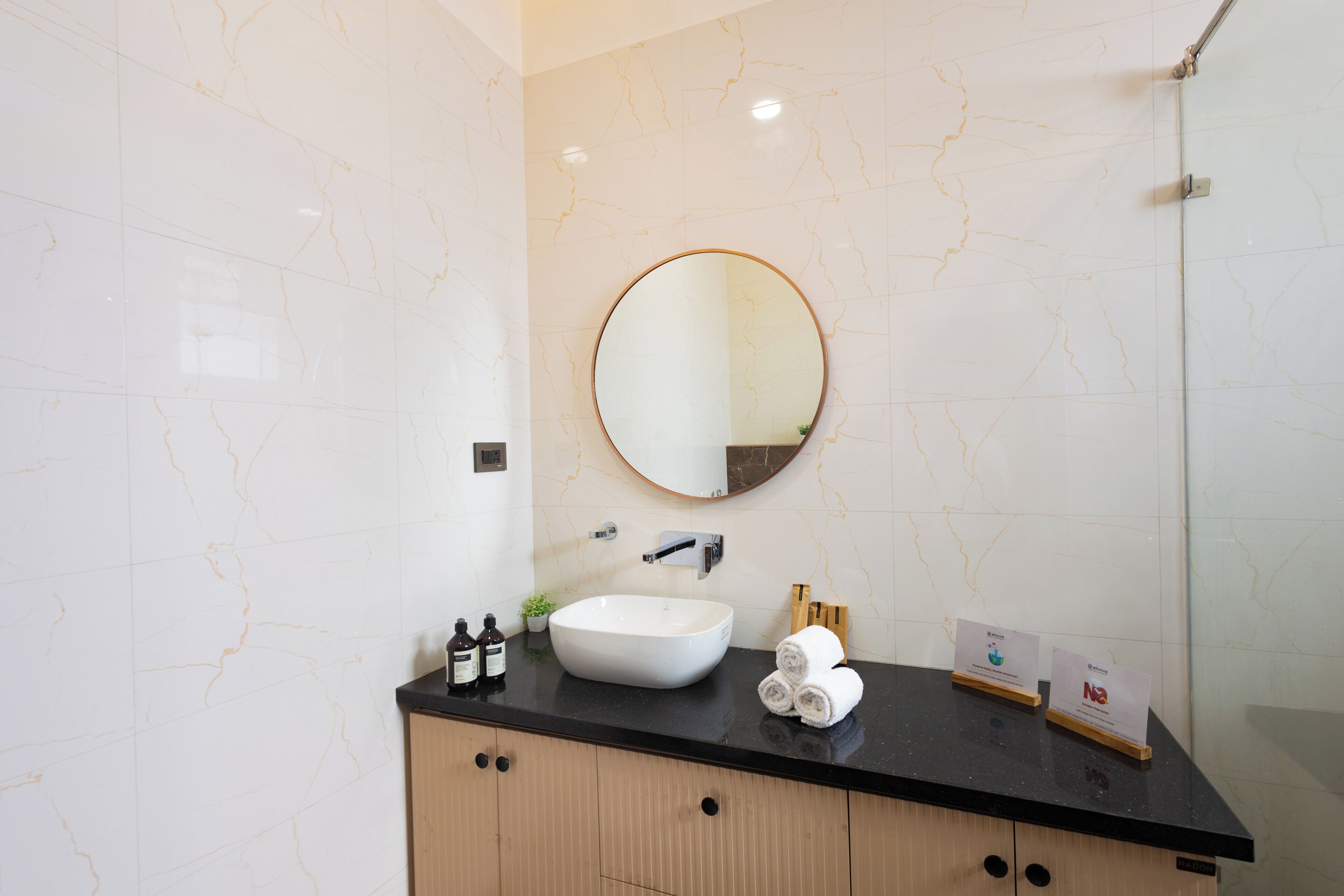 Un baño con pared de mármol blanco, espejo redondo, encimera negra, lavabo blanco y un mueble de madera.