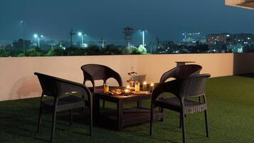 A rooftop terrace with a table set for two, surrounded by wicker chairs, and a cityscape view in the background.