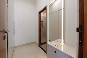 A hallway with a door leading to a bathroom, a mirror, and a small table with items on it. - ELIVAAS Lush Haven 4BHK Villa Pvt Pool (Jaipur)