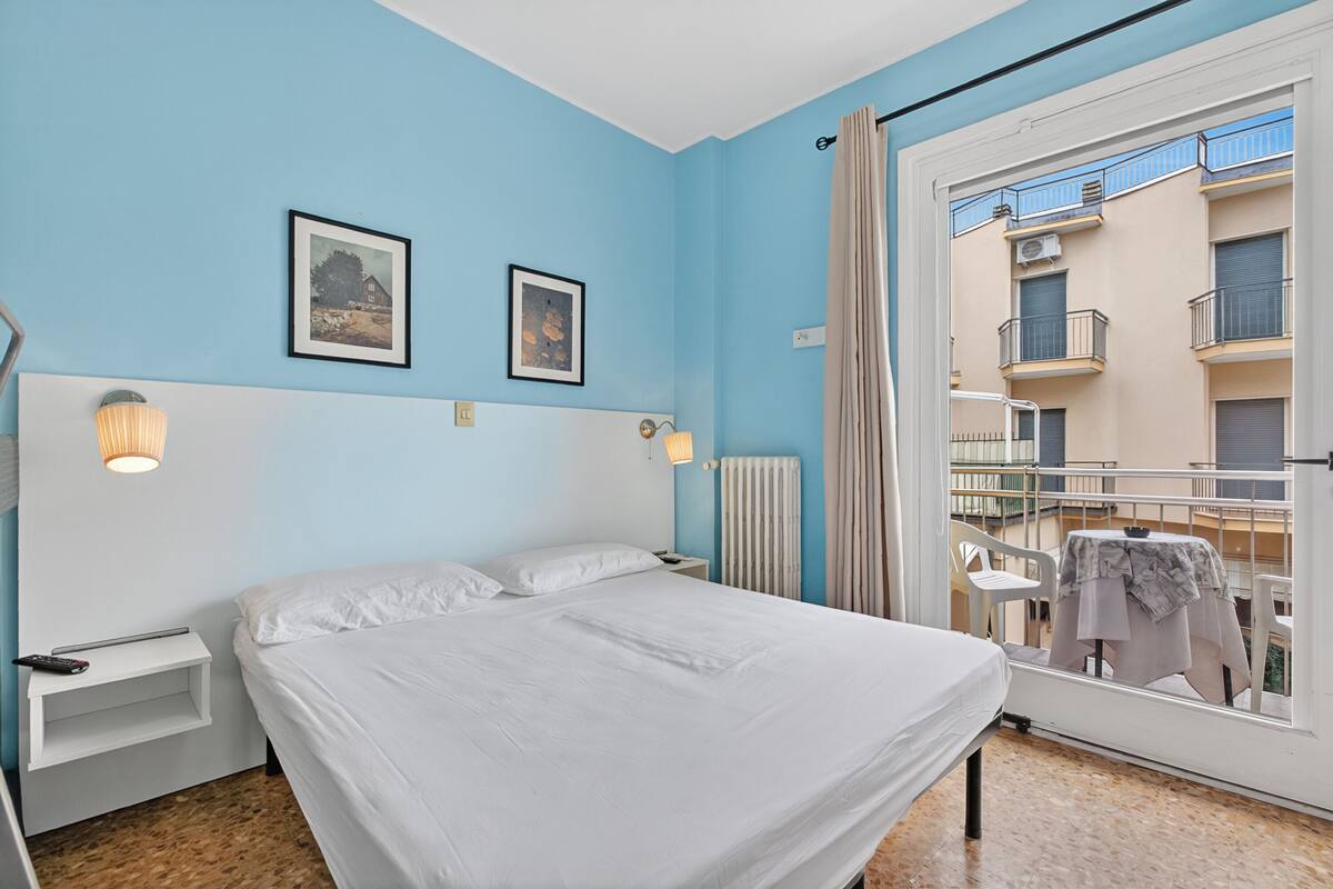 Een slaapkamer met een bed, nachtkastjes, wandlampen en een raam met uitzicht op een balkon.