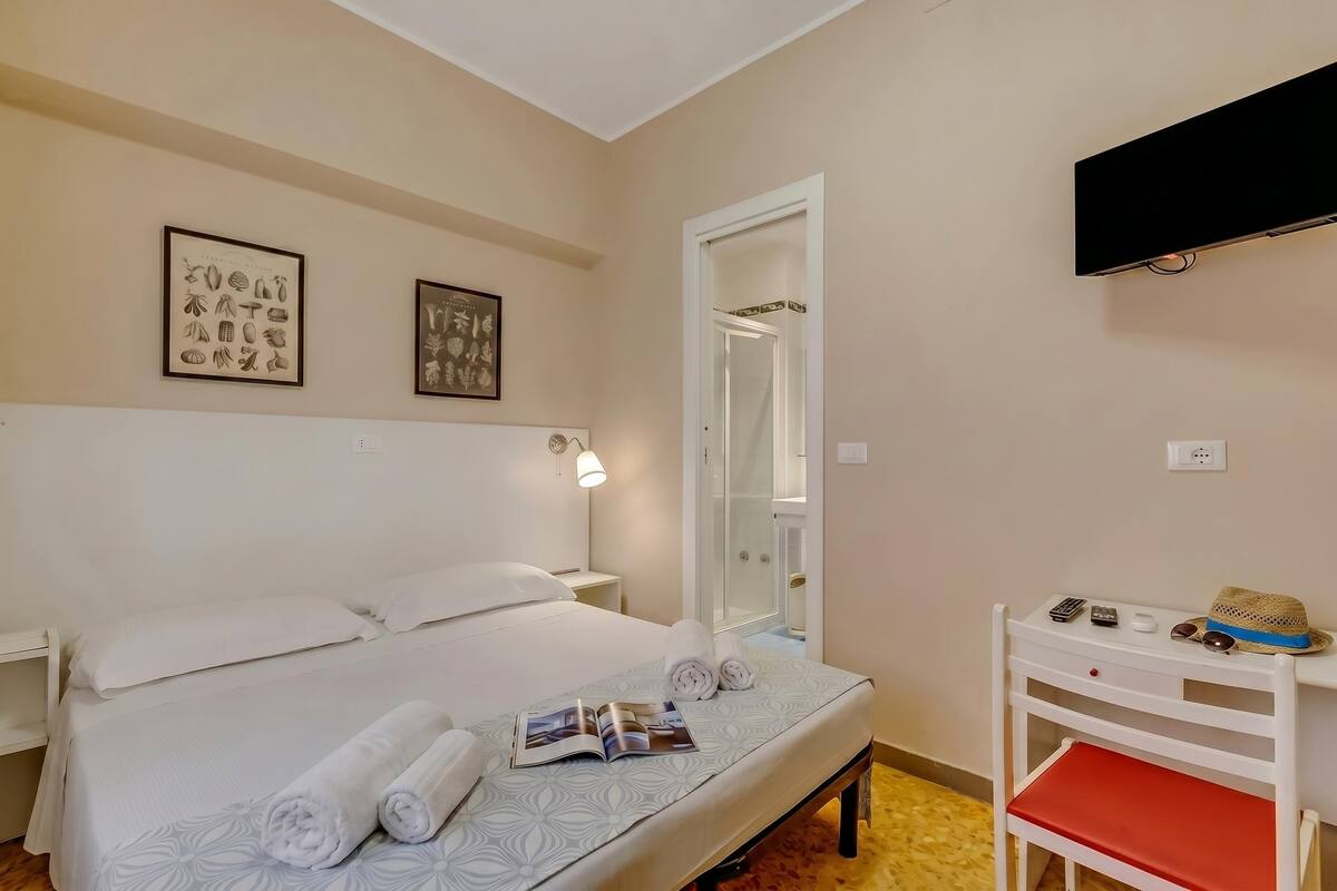 A hotel room with a bed, bedside tables, a TV, a mirror, and a small desk with a hat and sunglasses.