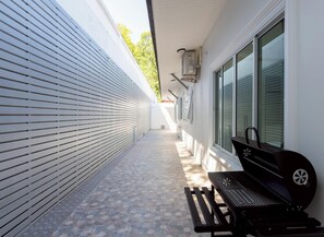 Ein schmaler Außenkorridor mit Grill und Bank, eine weiße Wand mit horizontalen Rollläden und einem Fenster mit Jalousien.
