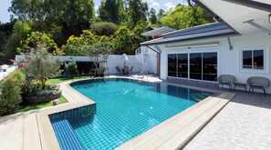 Ein Pool mit gefliestem Rand, eine überdachte Terrasse mit Sitzgelegenheiten und ein weißes Gebäude mit großen Fenstern.