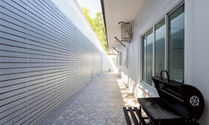 Ein schmaler Außenkorridor mit Grill und Bank, eine weiße Wand mit horizontalen Rollläden und einem Fenster mit Jalousien.