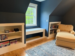 A room with a sloped ceiling, a window, built-in shelving with books, a beige armchair, and a blue rug.