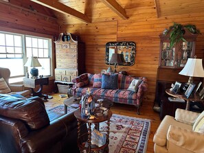 Ein gemütliches Blockhaushaus-Interieur mit Holzbalkendecke, einem karierten Sofa, einem Ledersessel, einer Holzkommode und einem Spiegel mit dekorativem Rahmen.