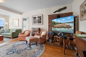 A living room with a fireplace, a bookshelf, a green sofa, and a large TV screen displaying a scenic view. - One Atlantic- | 5BR harbor view Maine home, close to town, deck (Boothbay Harbor)
