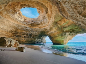 A natural rock formation with a circular opening, a sandy beach, and clear blue water. - Boho Sea by BeCherish (Albufeira)