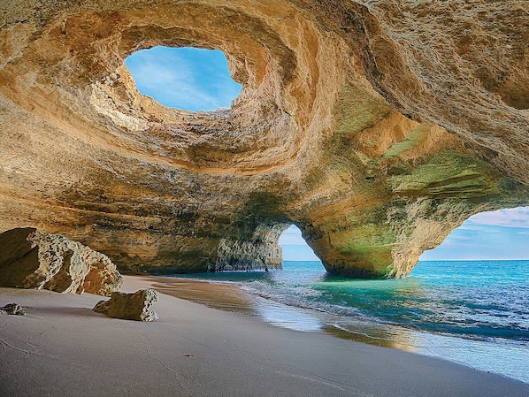 A natural rock formation with a circular opening, a sandy beach, and clear blue water. - Boho Sea by BeCherish (Albufeira)