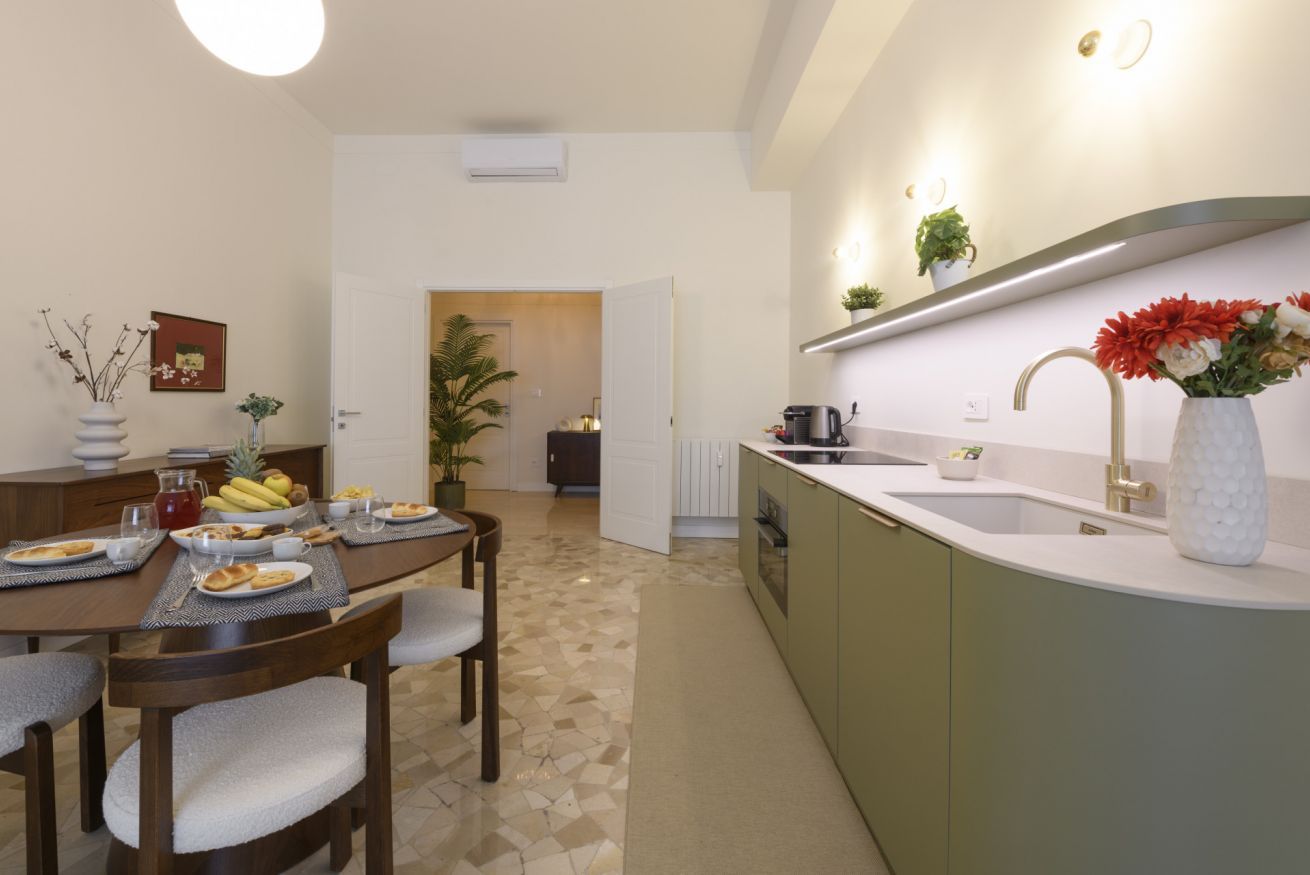 A modern kitchen with a dining area, featuring a central island, a built-in oven, and a sink with a gold faucet.
