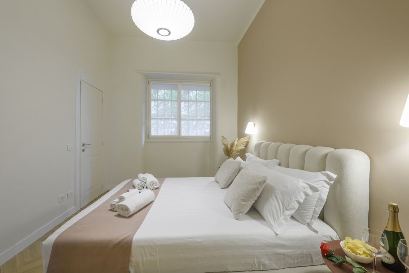 A hotel room with a bed, a window, a door, and a nightstand with a bottle and glasses.