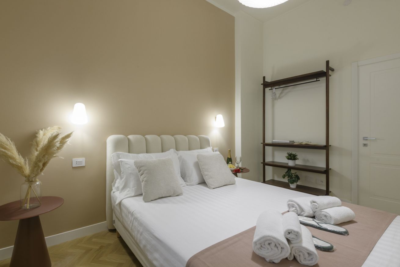 A hotel room with a bed, bedside lamps, a nightstand, a mirror, and a shelf with toiletries.