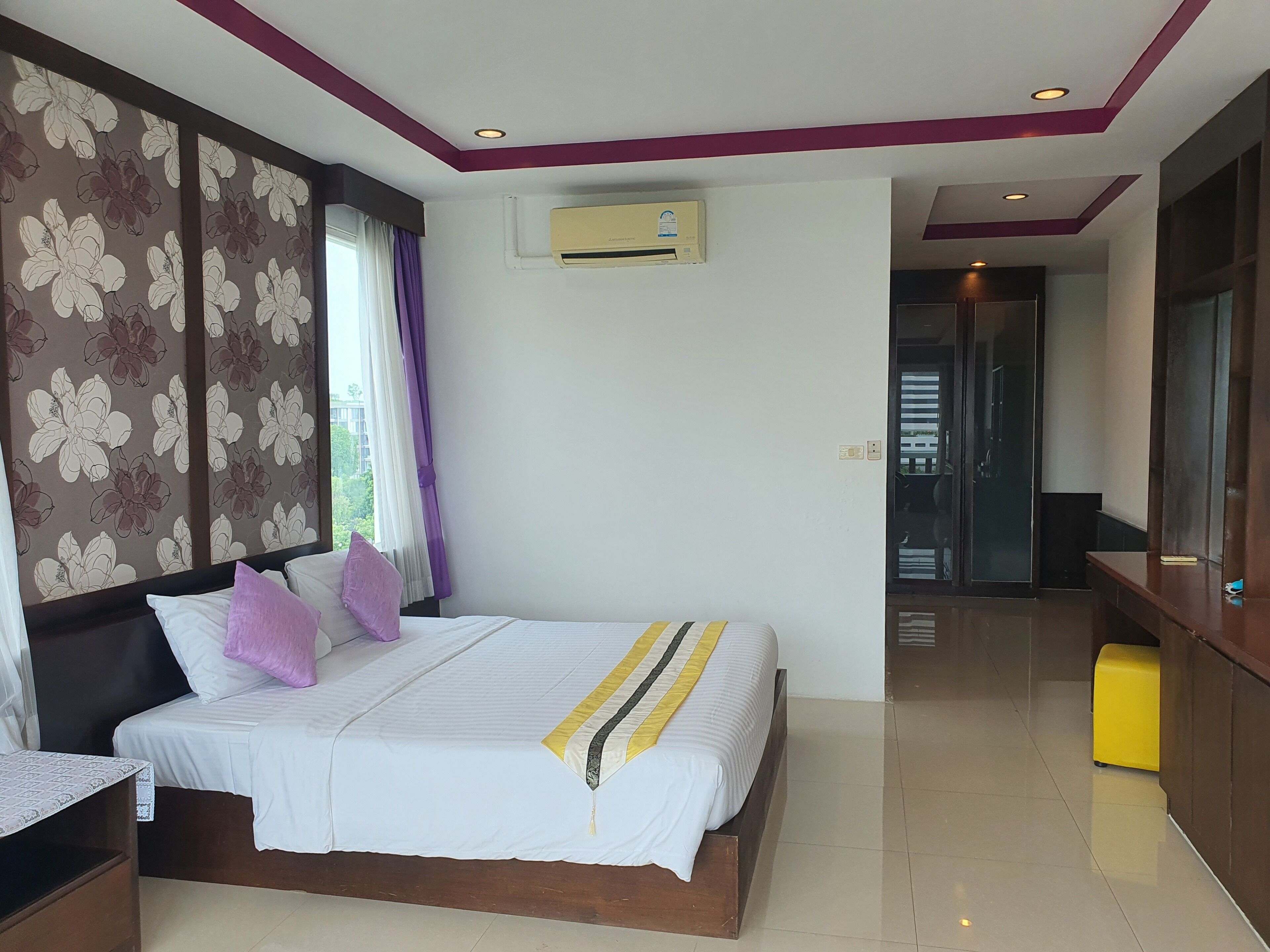 A hotel room with a bed, a nightstand, a desk, an air conditioning unit, and a view of the outside.