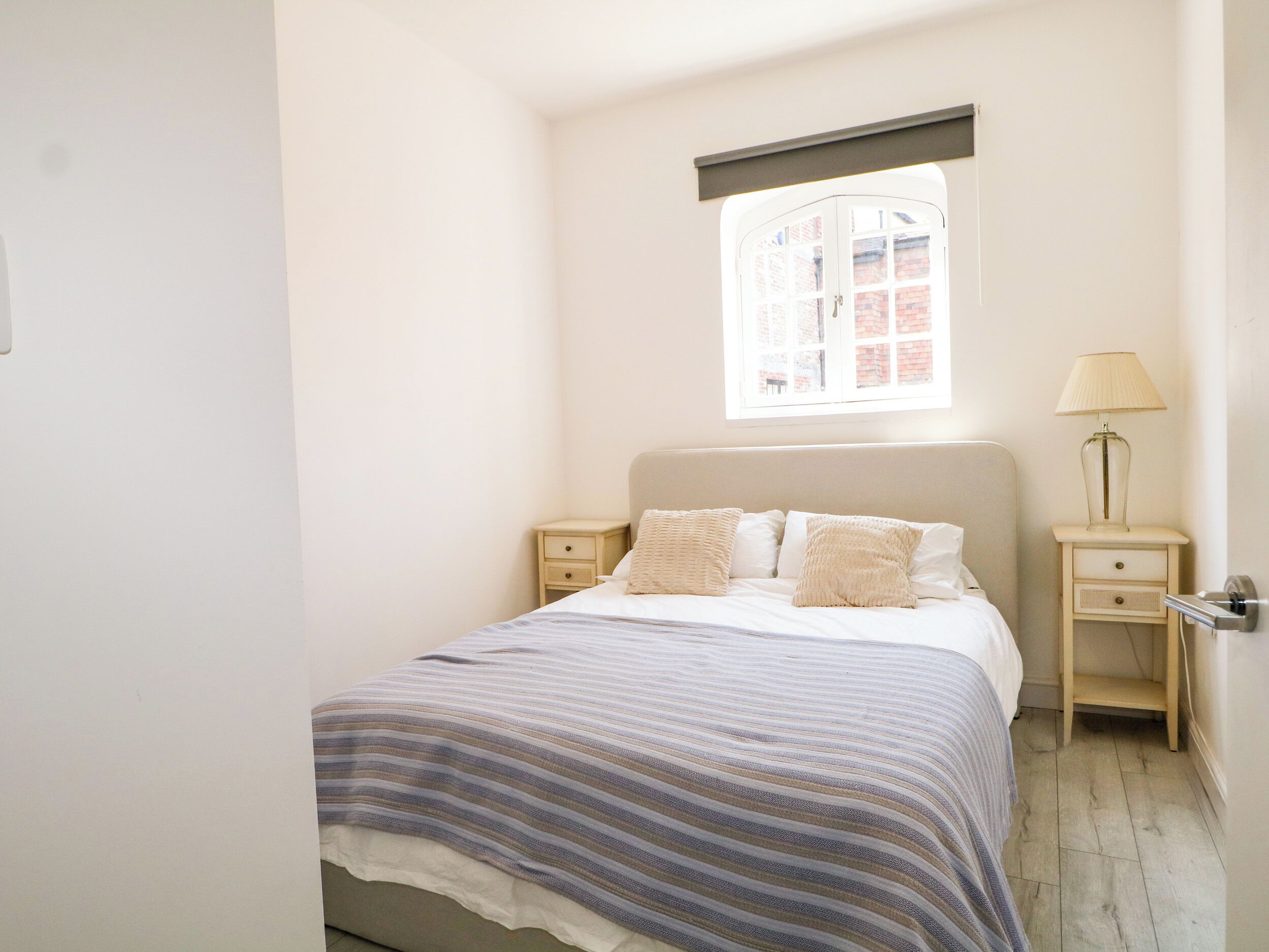 Une chambre à coucher avec un lit simple, une table de chevet, une lampe et une fenêtre donnant sur un immeuble en briques.