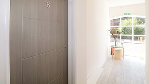 Een gang met een gesloten deur met het nummer 6, die leidt naar een lichte kamer met een raam en een glazen tafel met een vaas.