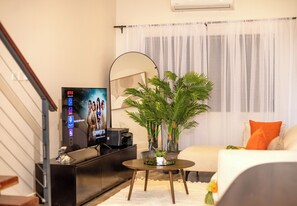 Ein modernes Wohnzimmer mit einem Flachbildfernseher, einem runden Couchtisch, einer Zimmerpflanze, einem Sofa mit orangefarbenem Kissen und einer Treppe.