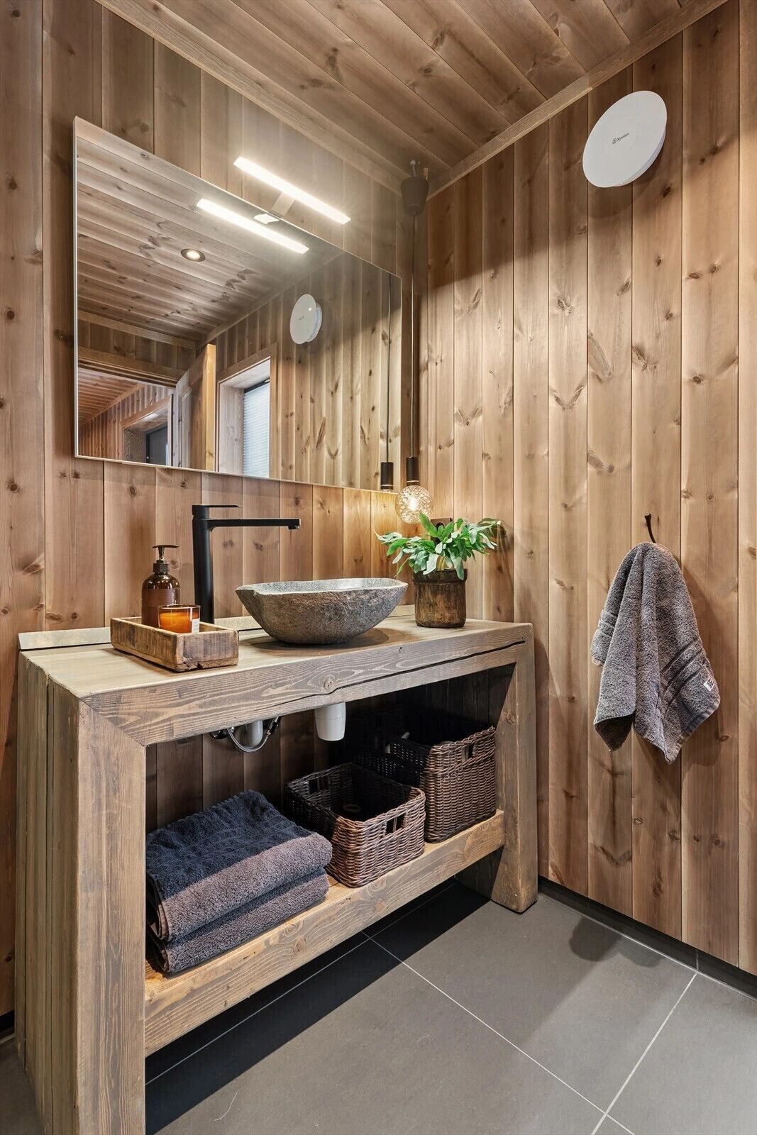 A wooden bathroom with a large mirror, a stone basin, a potted plant, a towel rack, and a wooden vanity.