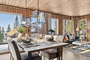 A modern interior with a wooden ceiling, large windows, a dining area, and a seating area with a view of trees.