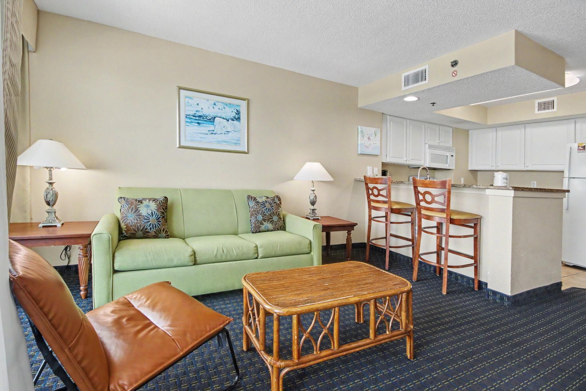 A hotel room with a green sofa, a wooden coffee table, a dining table with chairs, a kitchen area, and a framed picture on the wall.