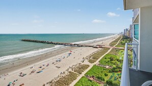 A beach view from a balcony overlooking a sandy beach with umbrellas, a pier extending into the water, and adjacent buildings. - Oceanfront 1BR Suite + Lazy River & Hot Tub (Myrtle Beach)