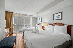 Ein Hotelzimmer mit zwei Betten, einem Holz-Kopfteil, einer Bank, einem Schminktisch und einem großen Fenster mit Vorhängen.