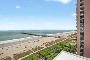 Blick vom Hotelbalkon auf einen Strand, mit einem Steg, der sich ins Meer erstreckt, sowie Sand und Grün im Vordergrund.