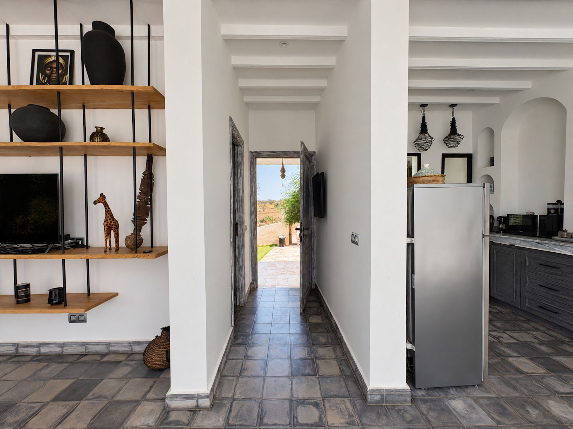A modern kitchen with a refrigerator, microwave, and cabinets. There is a hallway leading to a patio with a view of the landscape.